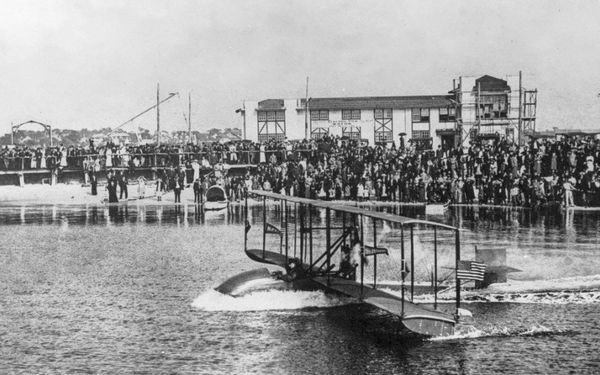 Black-and-white photo of a 1914 Benoist flying boat speeding across Tampa Bay while a large crowd watches from the shore