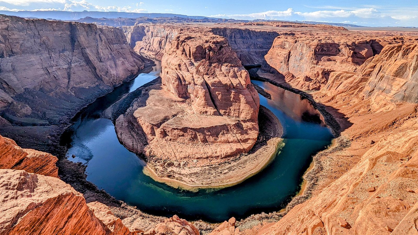 The Colorado River curving in a deep horseshoe-shaped bend around a tall red sandstone formation, seen from the canyon rim under a blue sky