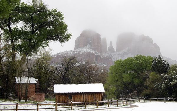 Snow covering the ground, a fence and two small historic cabins in front of red rock buttes at Sedona, with the tops of the rocks hidden in low winter clouds