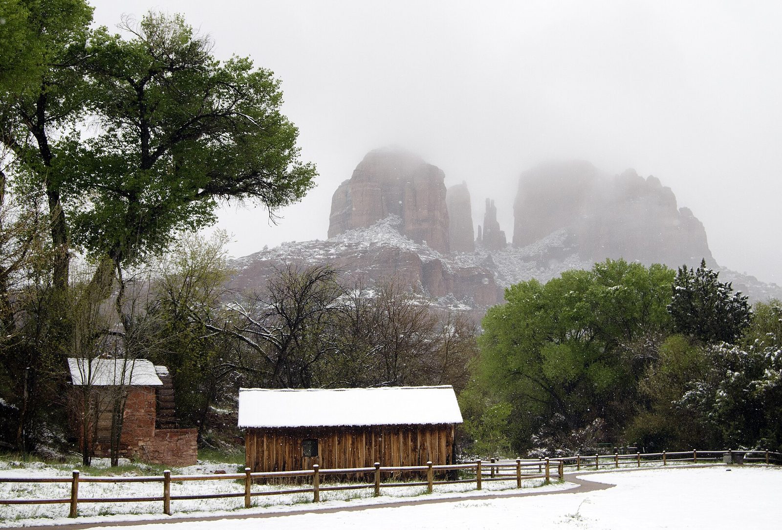 Snow covering the ground, a fence and two small historic cabins in front of red rock buttes at Sedona, with the tops of the rocks hidden in low winter clouds