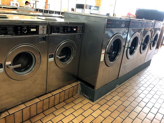 A row of stainless-steel front-loading washing machines in a self-service laundromat with tiled floors