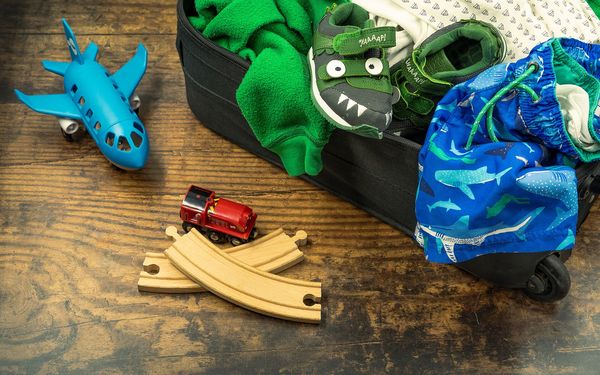 A child's black suitcase spilling over with a green fleece, dinosaur sneakers and shark-print swim shorts, beside a toy plane and a toy train on a wooden floor