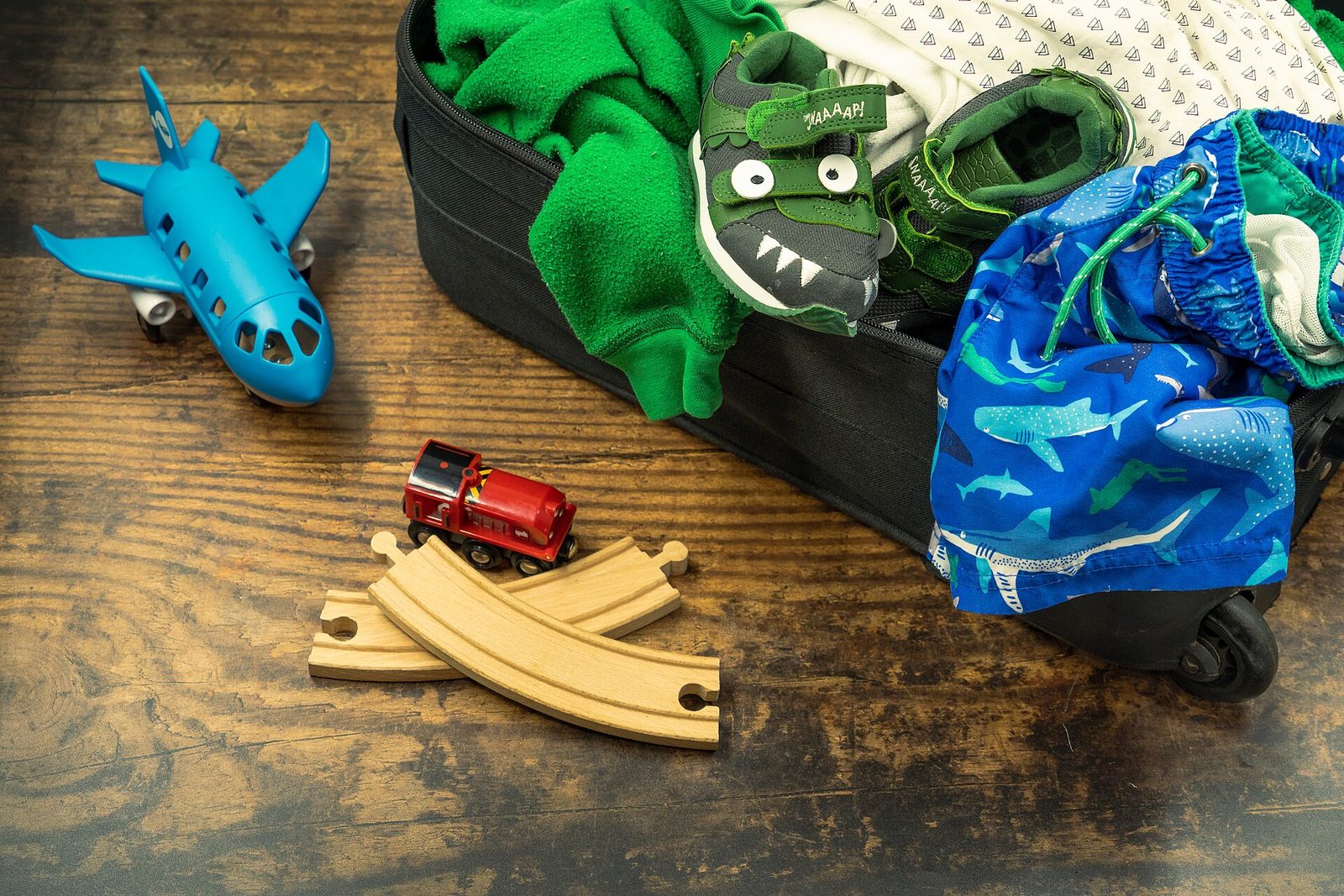 A child's black suitcase spilling over with a green fleece, dinosaur sneakers and shark-print swim shorts, beside a toy plane and a toy train on a wooden floor