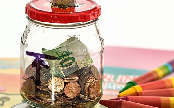 A glass jar used as a savings bank, filled with coins and a folded banknote, with a coin in the slot of its red lid and crayons on the table beside it