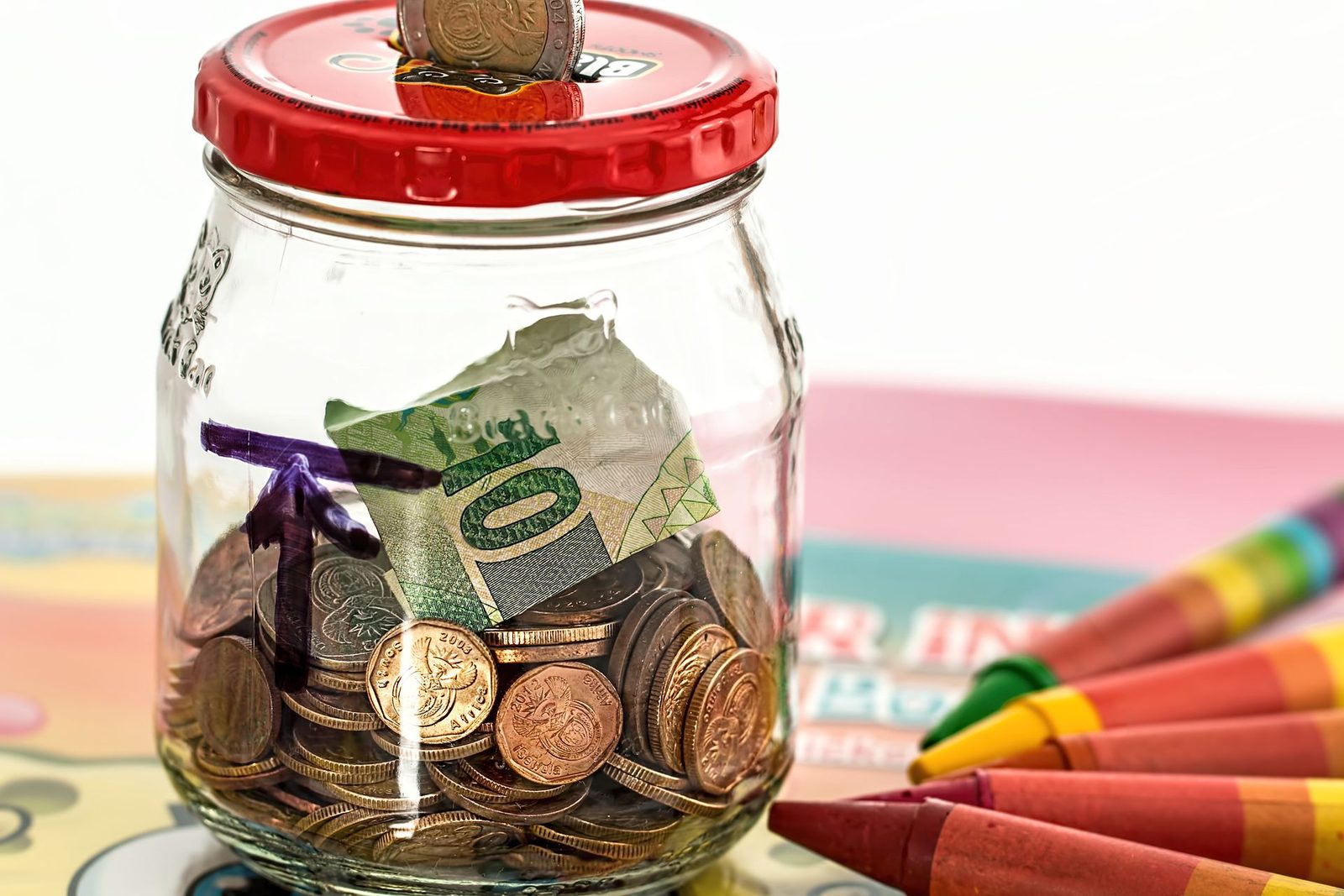 A glass jar used as a savings bank, filled with coins and a folded banknote, with a coin in the slot of its red lid and crayons on the table beside it