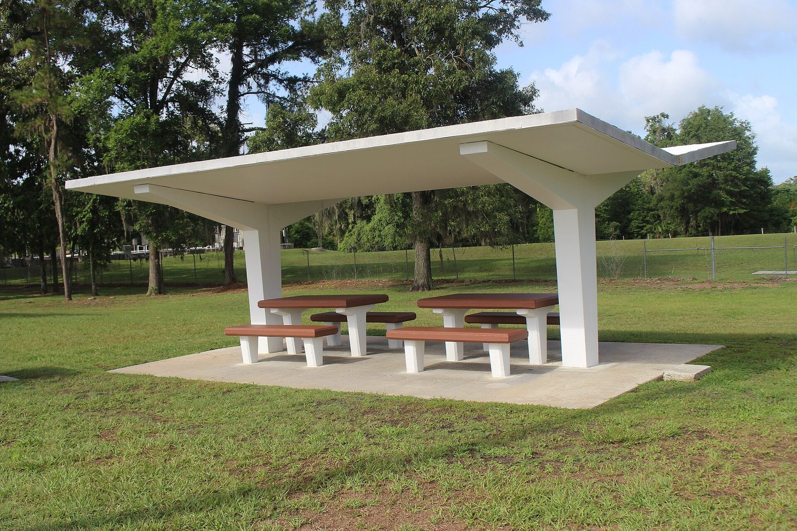 A covered picnic shelter with two brown picnic tables and benches on a grassy lawn at an interstate rest area, with tall trees behind