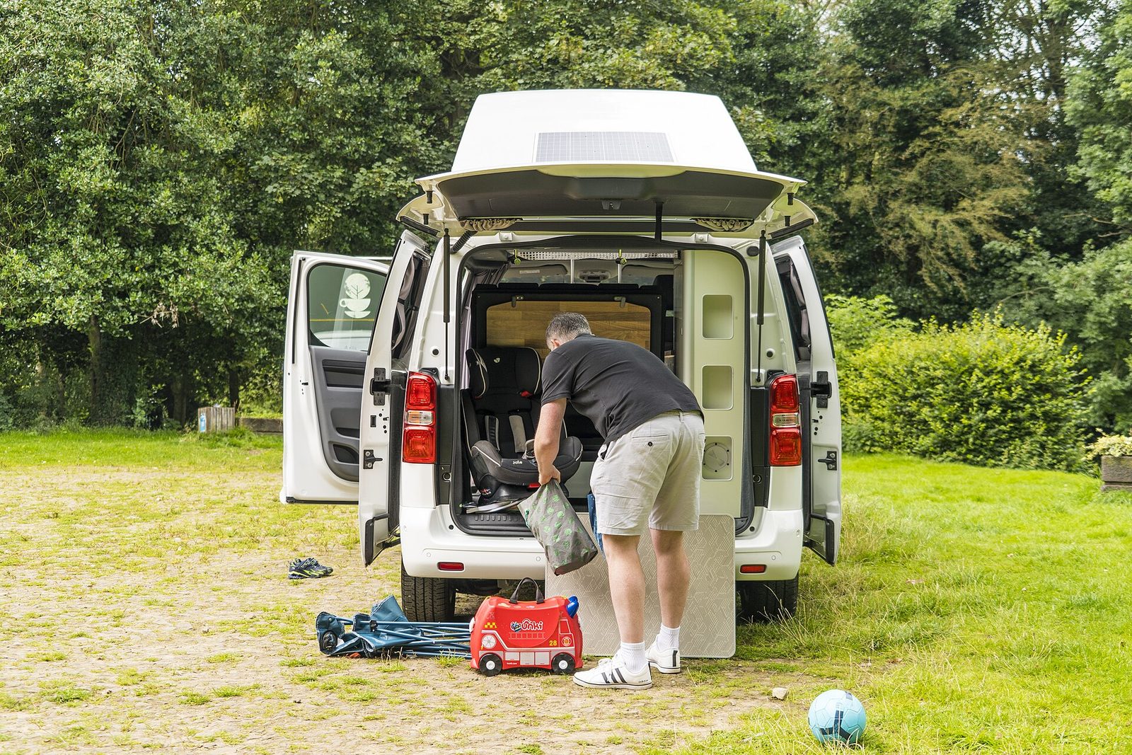 A man loads a bag into the open back of a white campervan with a child's car seat inside, with a small red ride-on suitcase, folded camping chairs and a ball on the grass