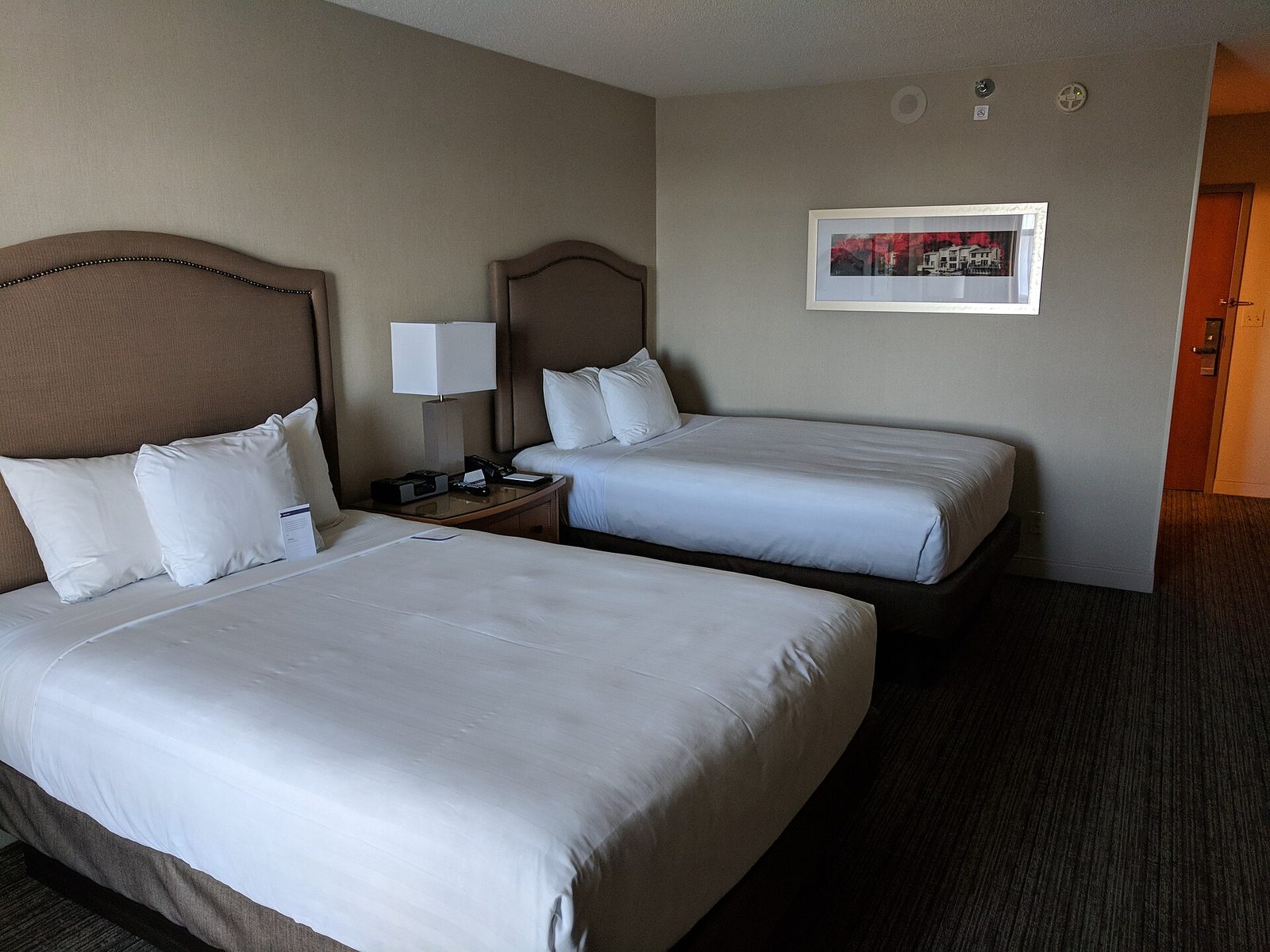 A plain hotel room with two queen beds, white bedding and a lamp on the nightstand between them