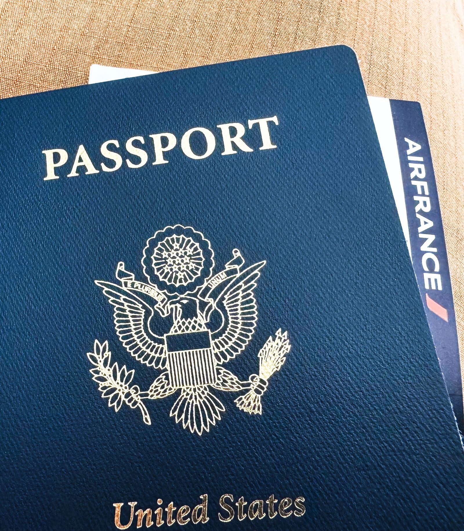 A navy-blue United States passport with a gold eagle seal, with an airline boarding pass tucked behind it, lying on a tan fabric surface