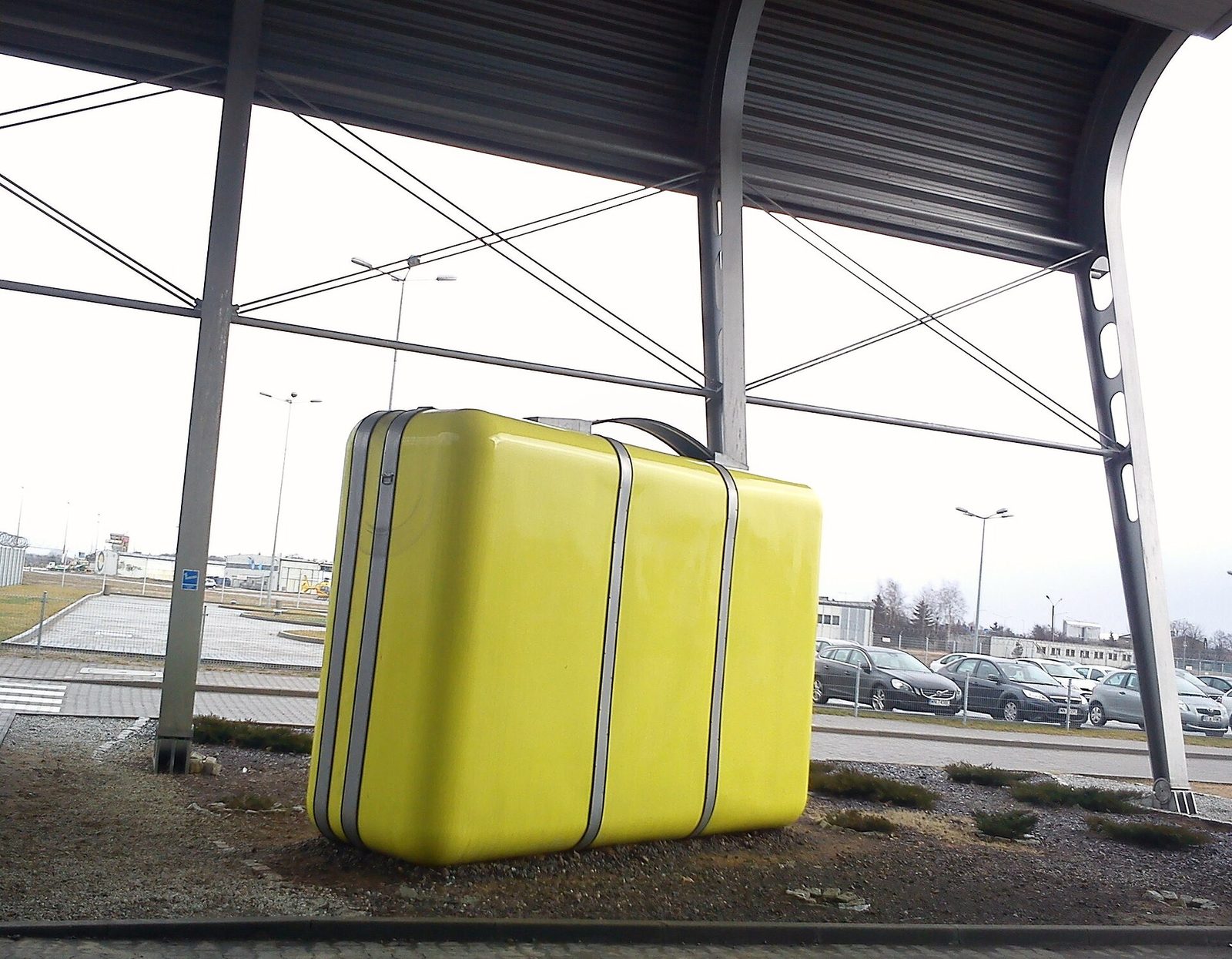 A giant yellow suitcase sculpture standing on gravel under a steel canopy outside an airport terminal, with a parking lot behind it