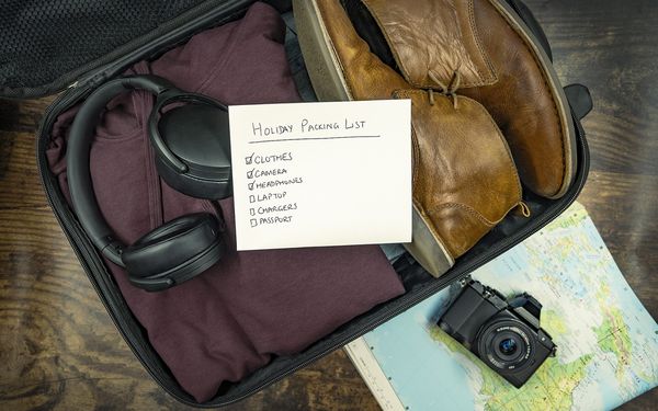 An open black suitcase with a folded maroon sweater, headphones and brown leather boots, with a handwritten holiday packing list on top and a camera resting on a map beside it