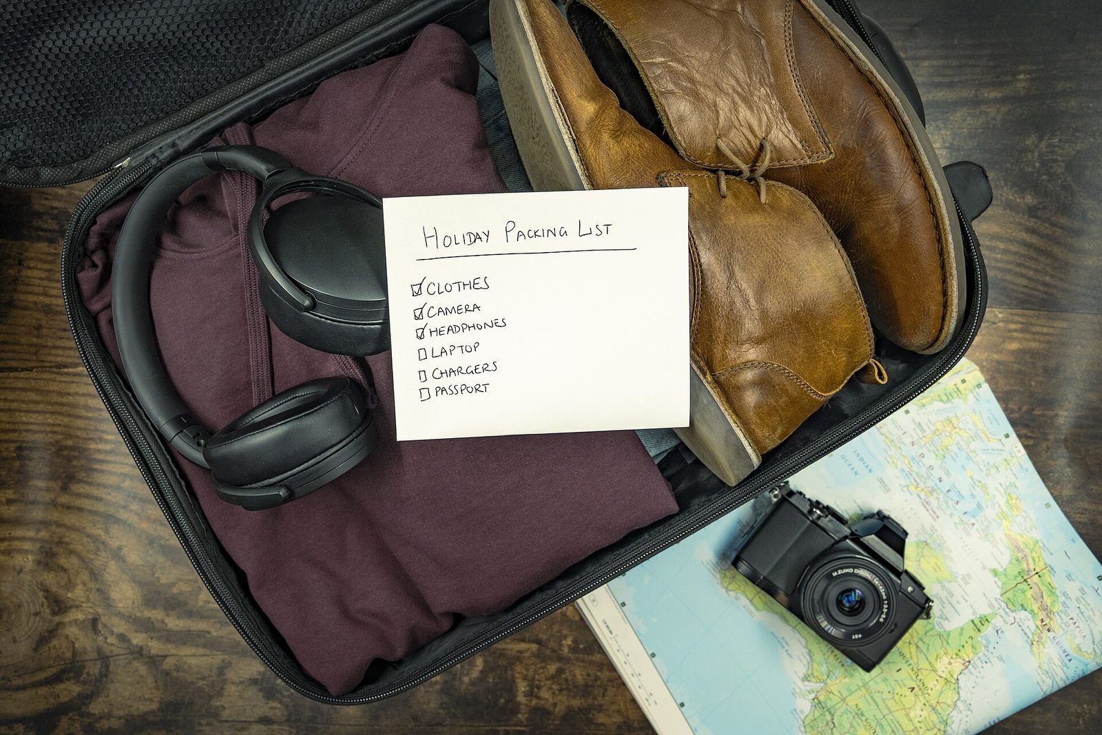 An open black suitcase with a folded maroon sweater, headphones and brown leather boots, with a handwritten holiday packing list on top and a camera resting on a map beside it