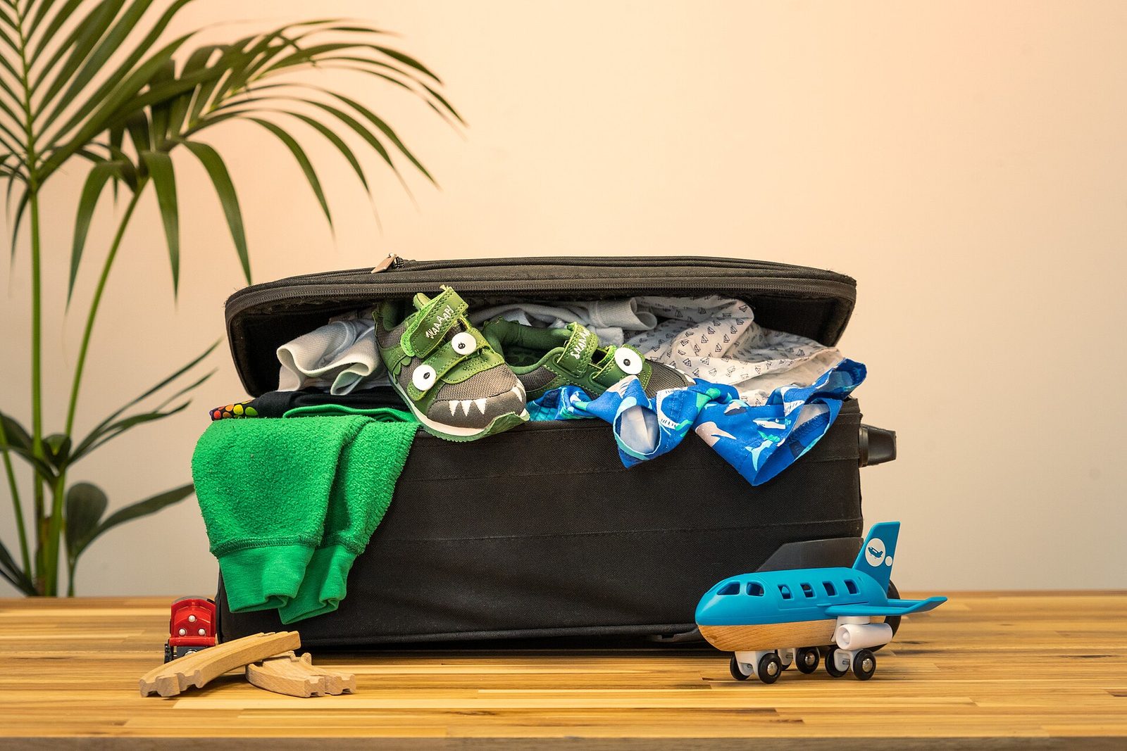 An open black suitcase overflowing with children's clothes and dinosaur sneakers, with a toy airplane and toy train beside it