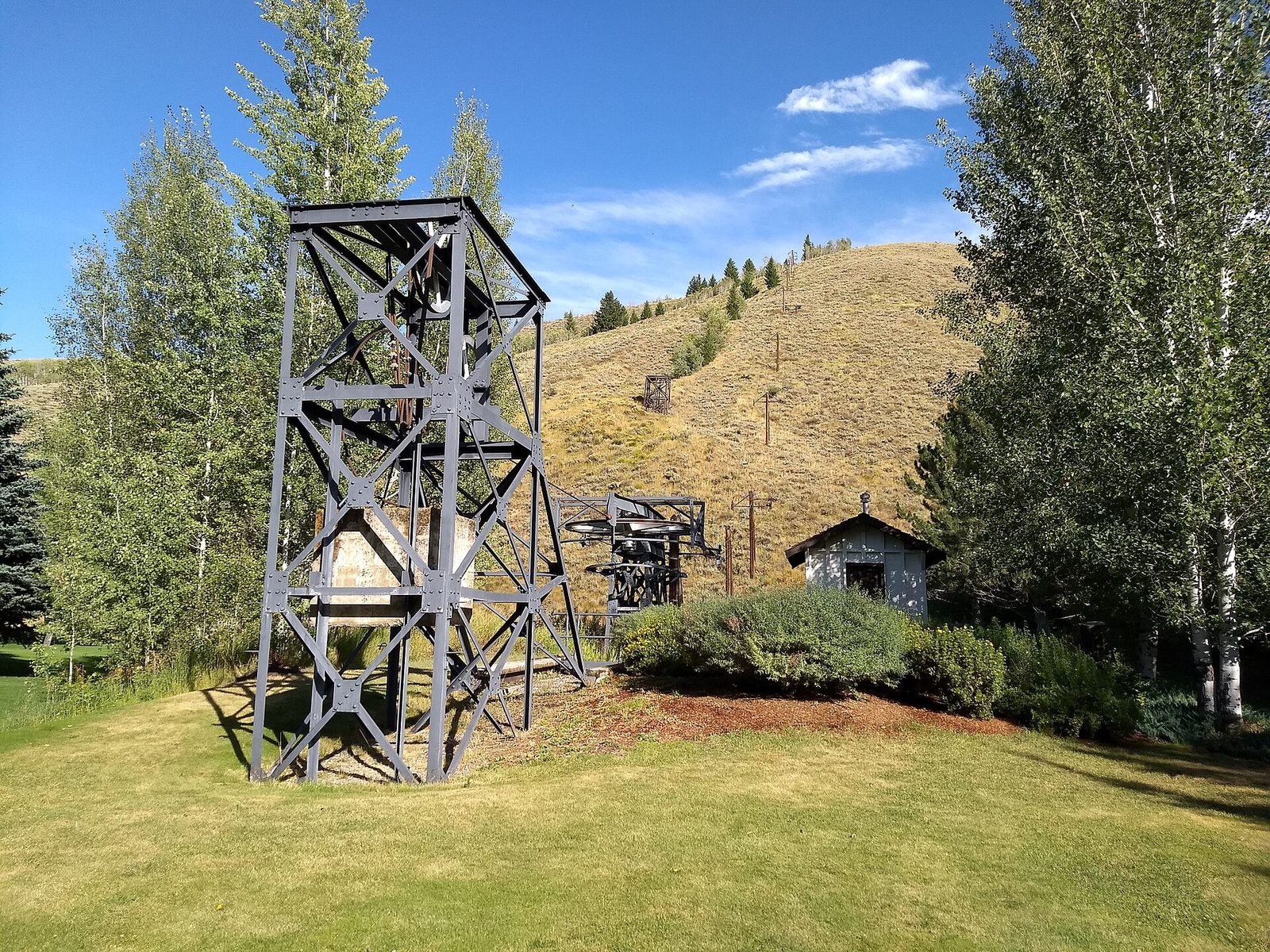 A tall dark steel lift tower and a large cable wheel stand on a green lawn in summer, with a line of old lift poles climbing a dry grassy hill behind them under a blue sky