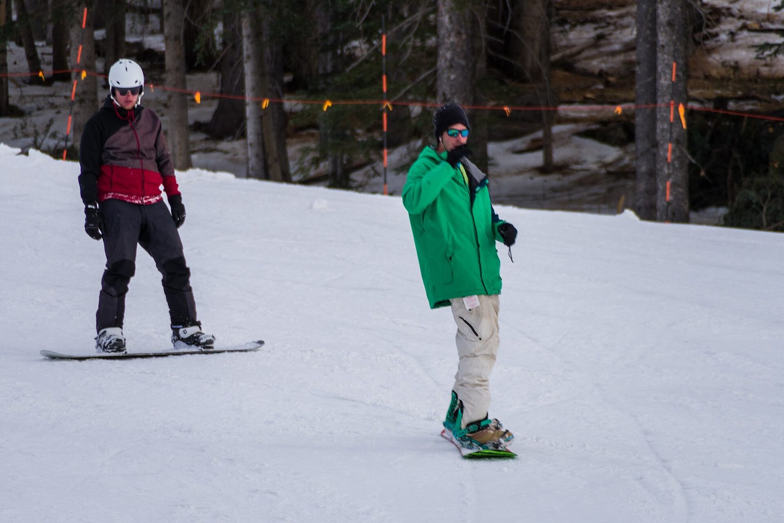 Two snowboarders in helmets, hats, goggles, waterproof jackets and snow pants ride down a groomed snowy slope lined with pine trees