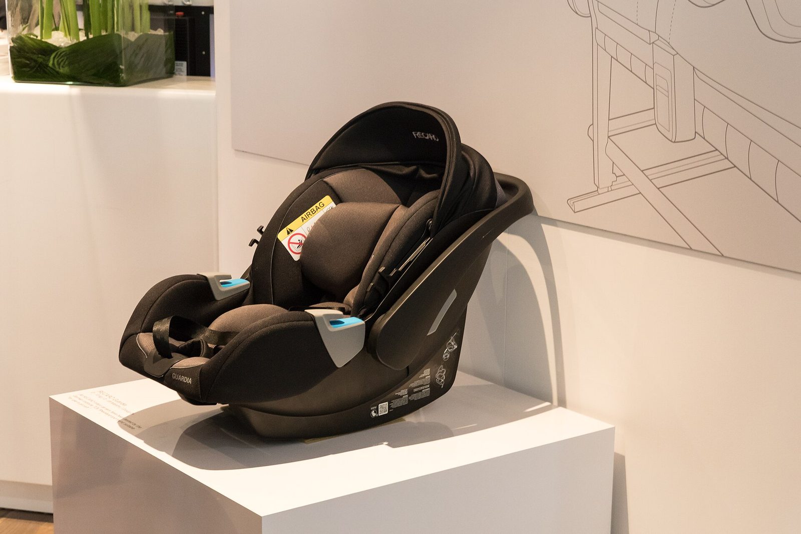 A black rear-facing infant car seat with a yellow airbag warning label, displayed on a white plinth at an aircraft-interiors trade show