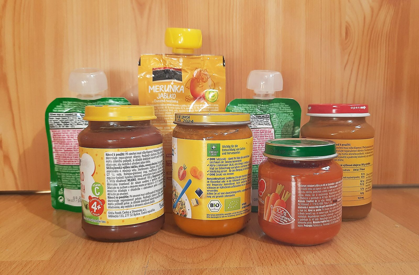Several jars of fruit and vegetable baby food and three squeeze pouches of fruit puree lined up on a wooden table