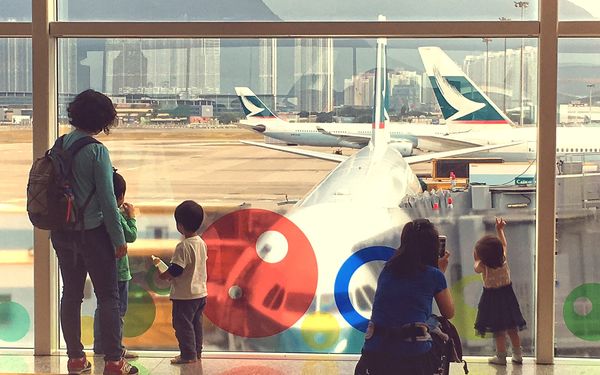 Two adults and three small children, seen from behind, watching parked airliners through a big airport terminal window