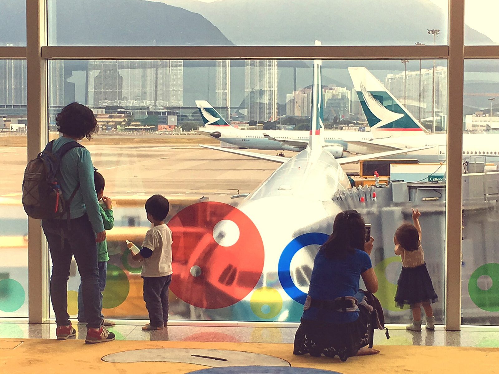 Two adults and three small children, seen from behind, watching parked airliners through a big airport terminal window