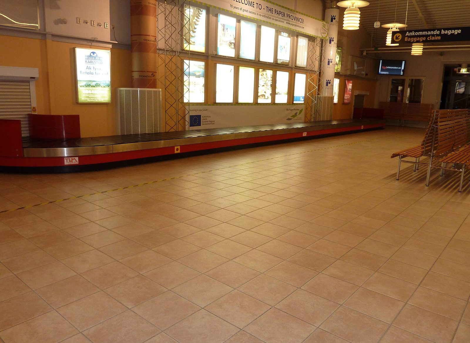 An empty airport baggage claim hall with a long red and steel luggage carousel, tiled floor, wooden benches and a Baggage claim sign