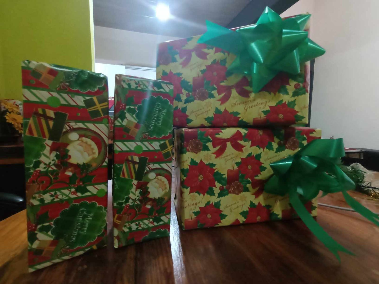 Four Christmas presents wrapped in red, green and gold holiday paper with green ribbon bows, stacked on a wooden table