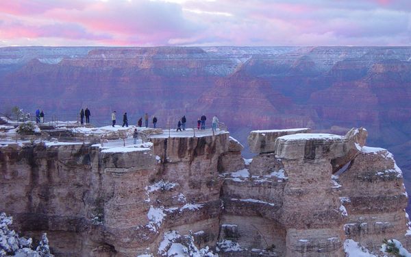 Visitors standing along the railed overlook at Mather Point at sunset, with fresh snow on the rim rocks and pink clouds over the canyon
