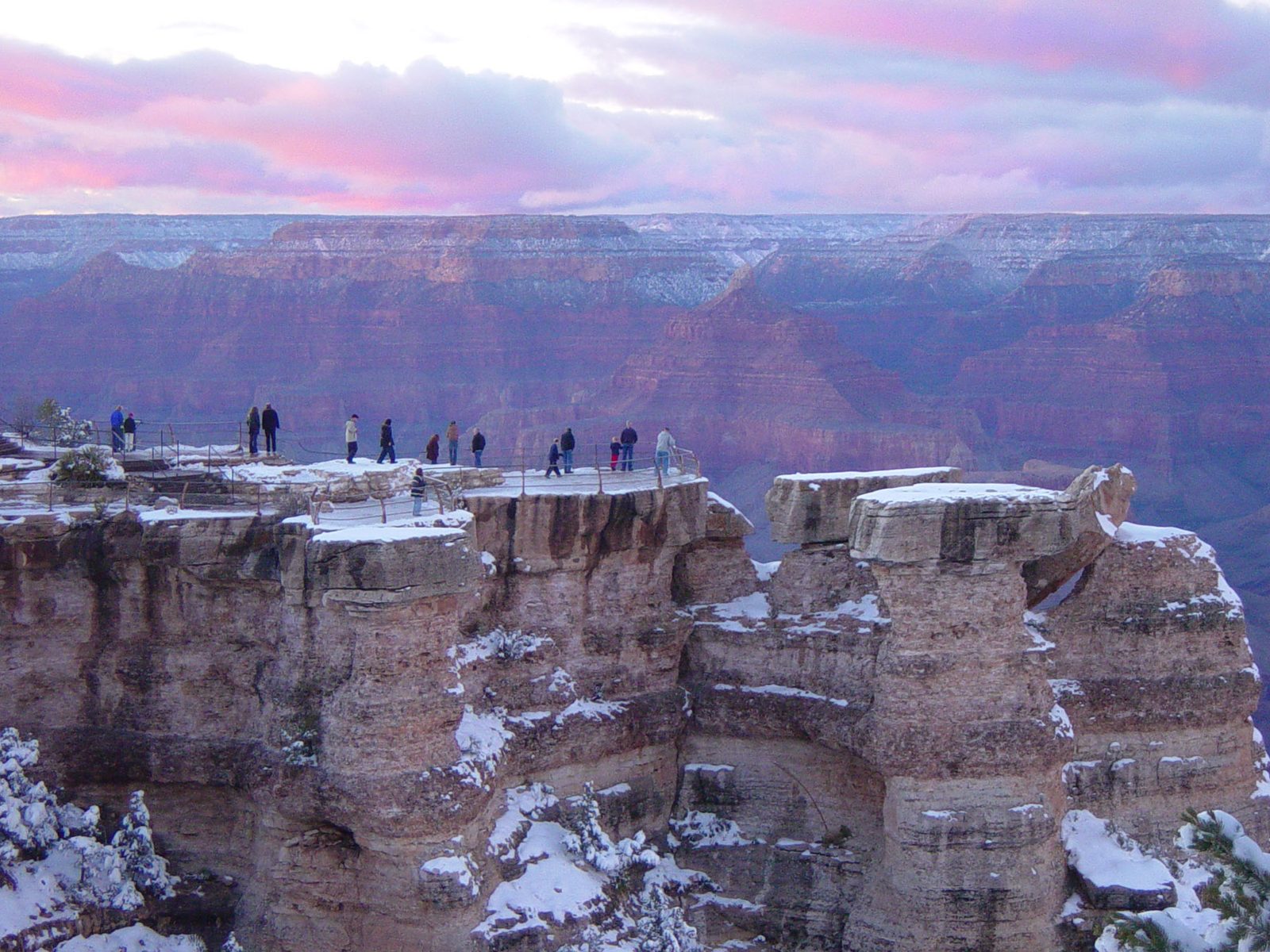 Visitors standing along the railed overlook at Mather Point at sunset, with fresh snow on the rim rocks and pink clouds over the canyon