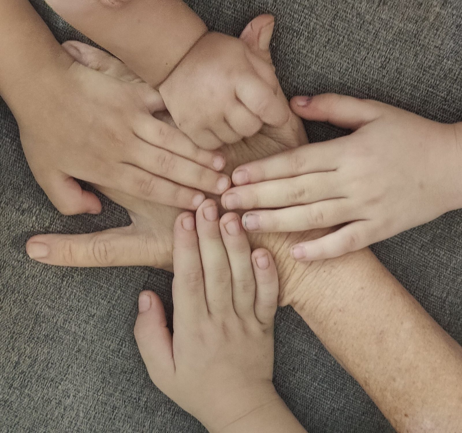Four small children's hands resting on top of an older woman's hand and forearm on a gray couch