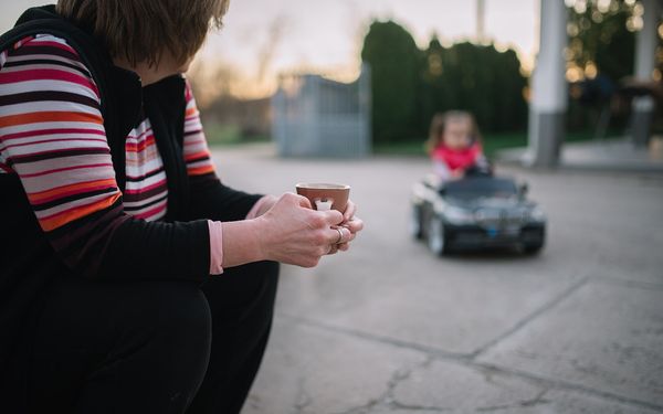 A grandparent holding a cup of coffee watches a small child drive a toy car on a driveway at sunset