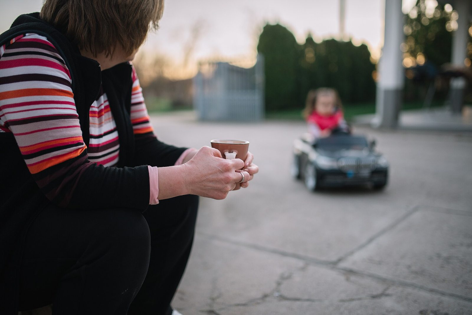 A grandparent holding a cup of coffee watches a small child drive a toy car on a driveway at sunset