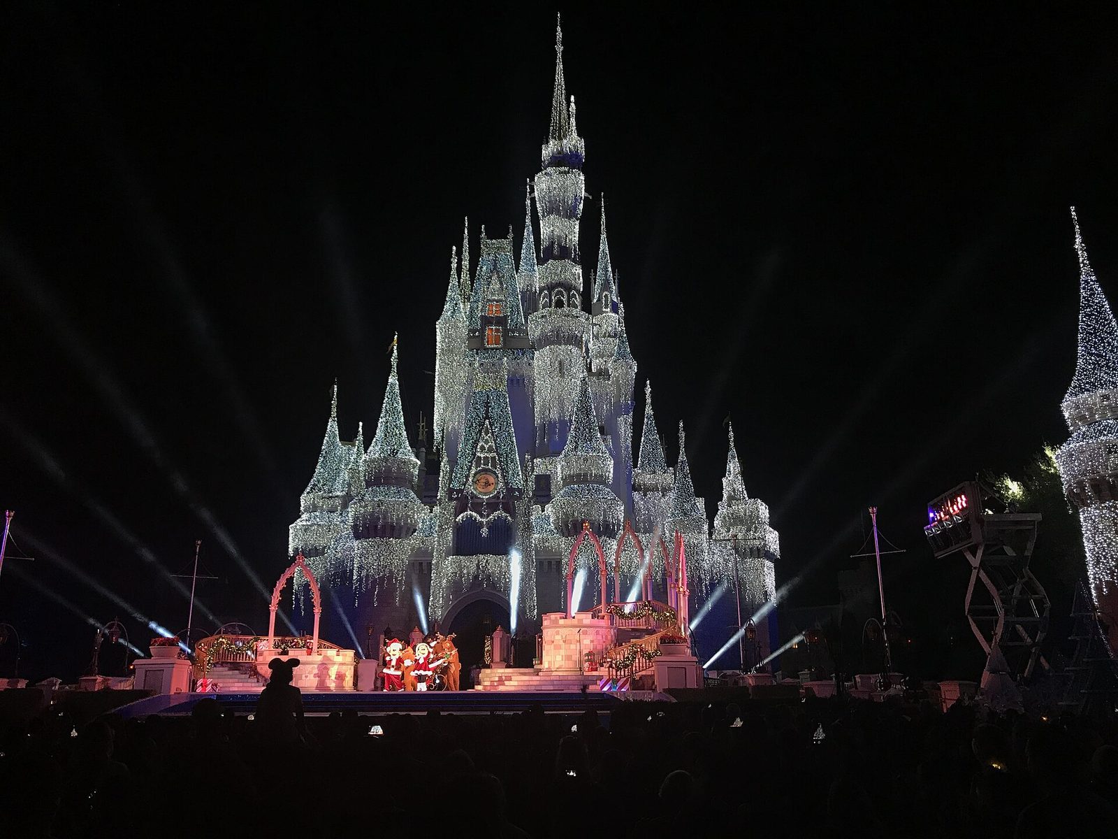 Cinderella Castle covered in white holiday lights at night, with Mickey and Minnie in red holiday outfits performing on the stage in front of a crowd