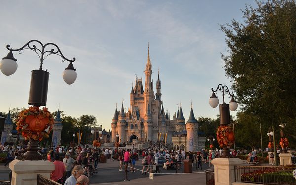 Cinderella Castle at Magic Kingdom in warm evening light, with Mickey-shaped pumpkin and fall-leaf decorations on the lamp posts and crowds on the plaza