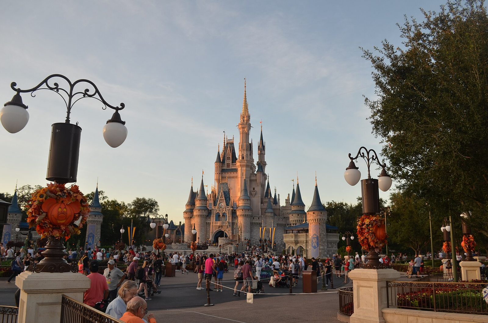 Cinderella Castle at Magic Kingdom in warm evening light, with Mickey-shaped pumpkin and fall-leaf decorations on the lamp posts and crowds on the plaza