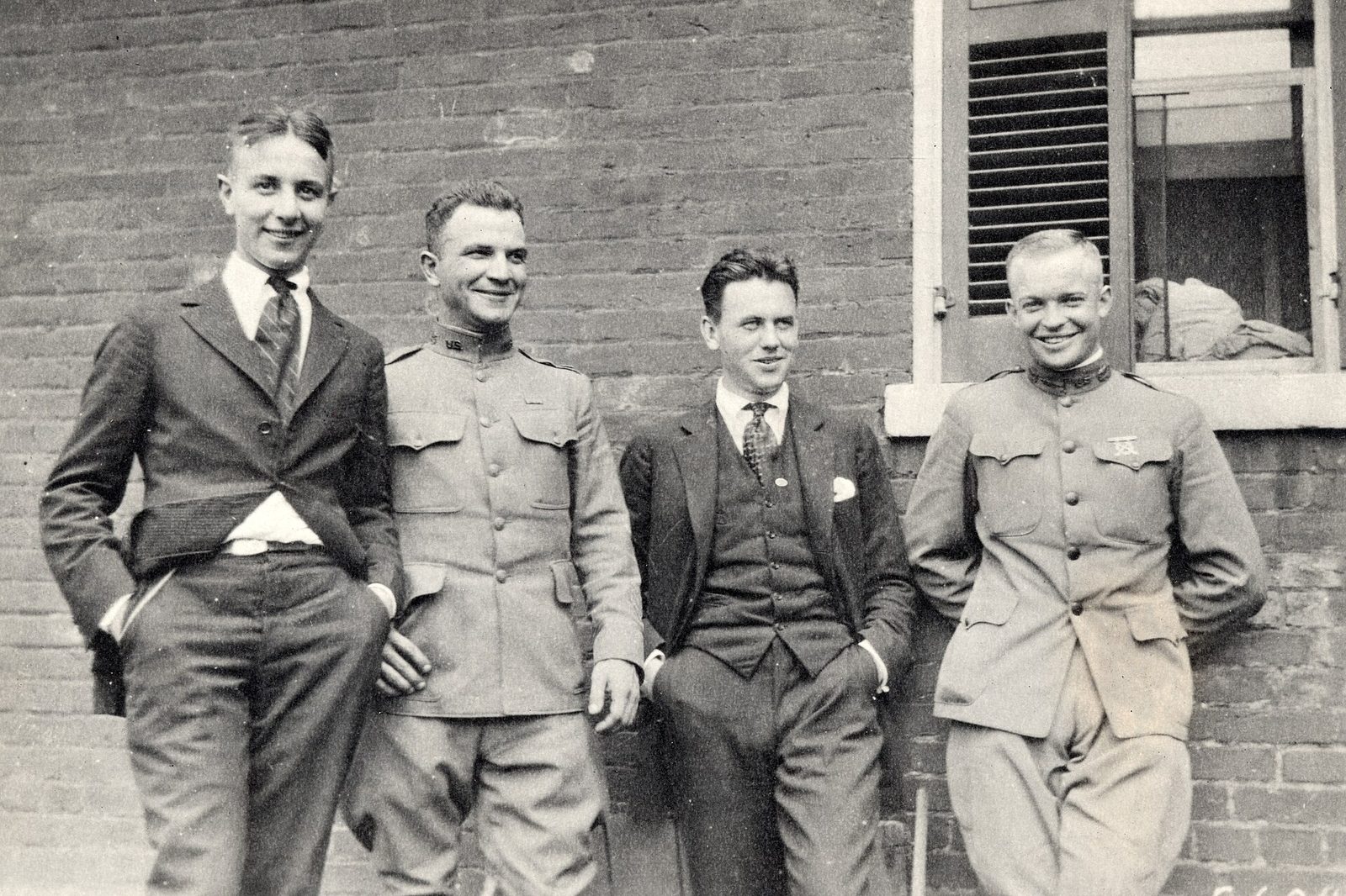 A black-and-white photo of four smiling young men leaning against a brick wall, two in early 1900s Army uniforms and two in suits, with Eisenhower on the far right
