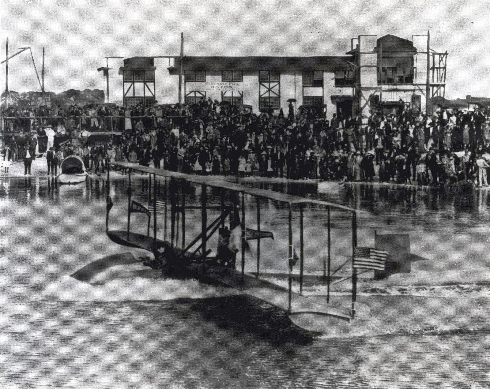 A black and white 1914 photograph of a small biplane flying boat with an American flag skimming across the water, with a large crowd watching from the shore in front of a waterfront building
