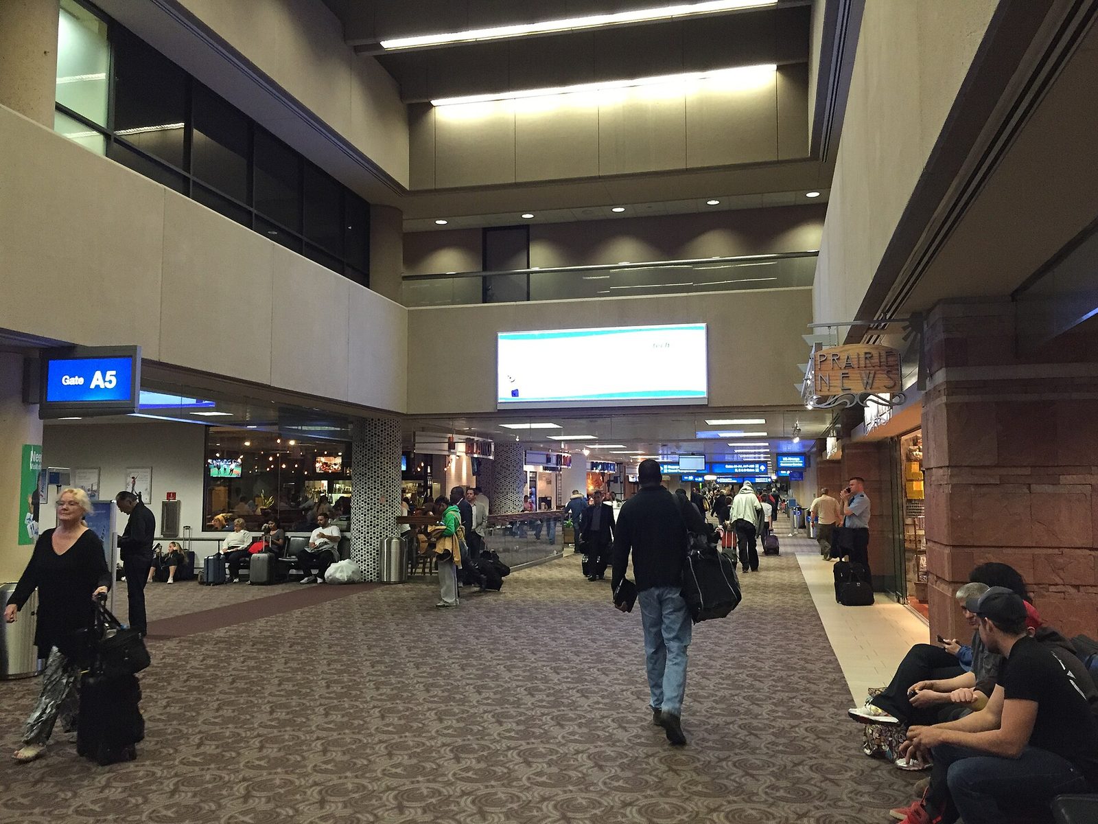 The inside of an airport terminal concourse with patterned carpet, a blue Gate A5 sign, shops on the right and travelers walking with rolling bags or sitting along the walls