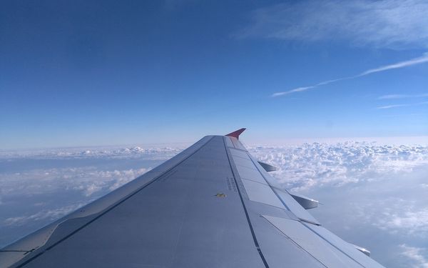 The view from an airplane window seat over a long silver wing with a red wingtip, a deep blue sky above and a floor of white puffy clouds below
