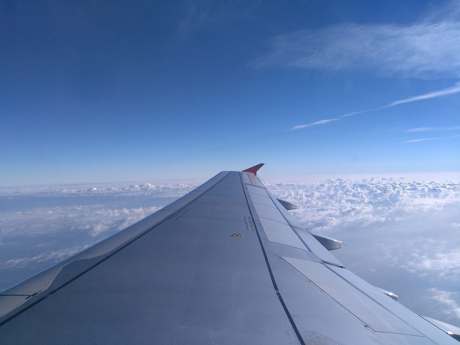 The view from an airplane window seat over a long silver wing with a red wingtip, a deep blue sky above and a floor of white puffy clouds below