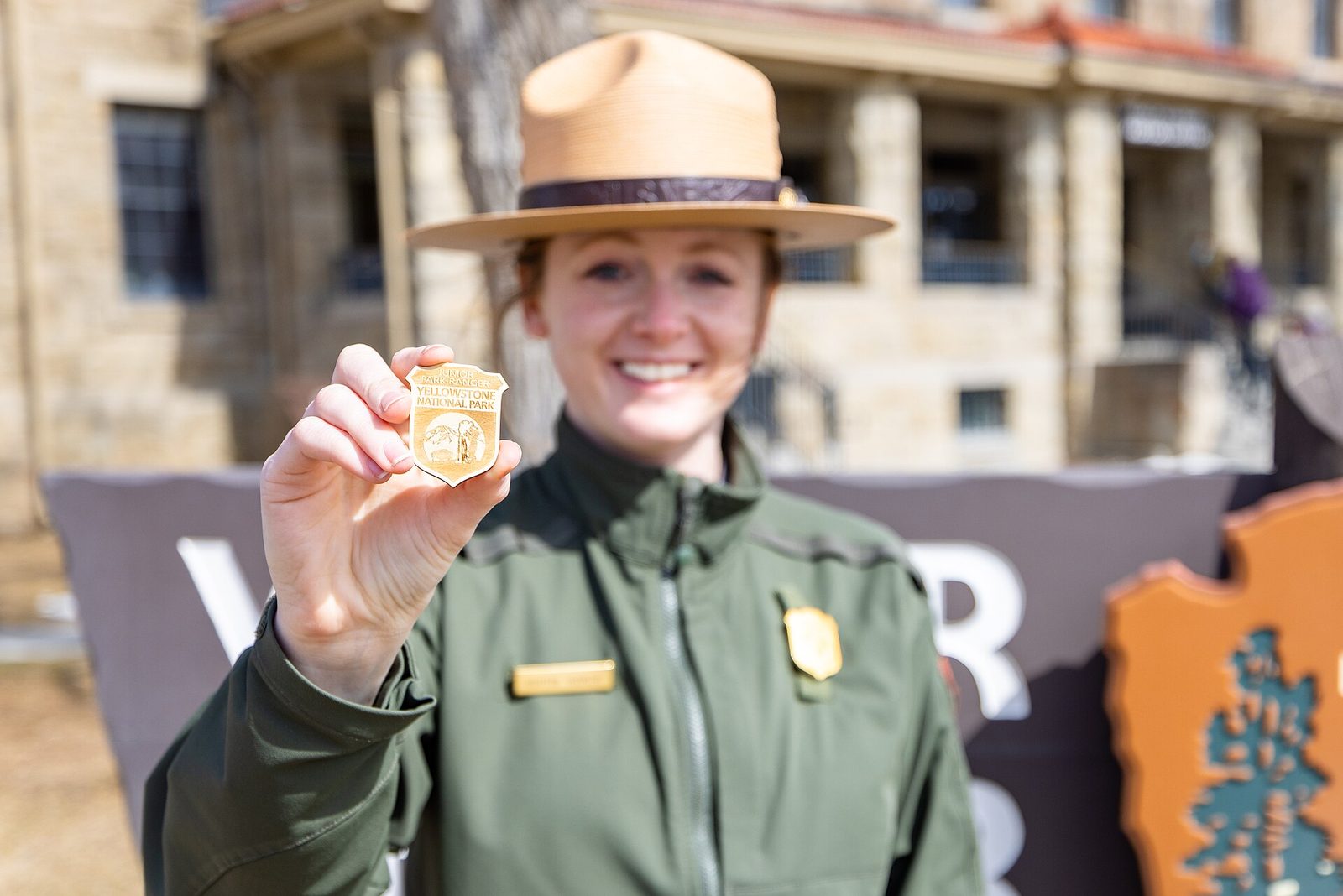 A smiling park ranger in a flat hat holds up a small gold Yellowstone Junior Ranger badge toward the camera
