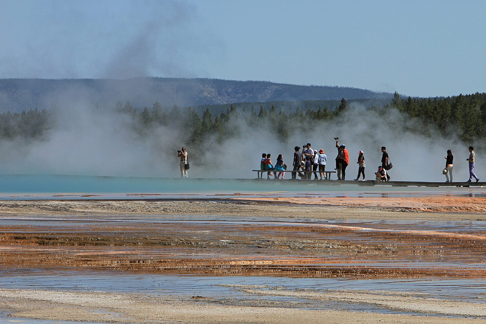 A group of visitors stands on a boardwalk at the edge of Grand Prismatic Spring as clouds of steam rise over the bright blue water and orange mineral flats