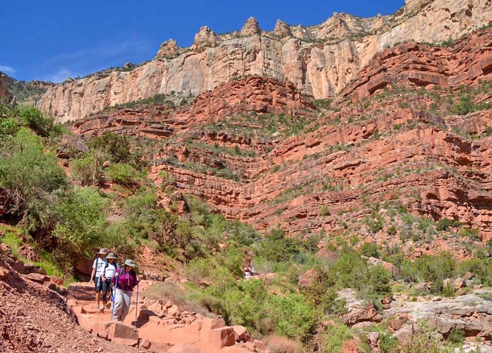 Three hikers with sun hats and trekking poles descending a rocky section of the Bright Angel Trail beneath red and tan canyon walls