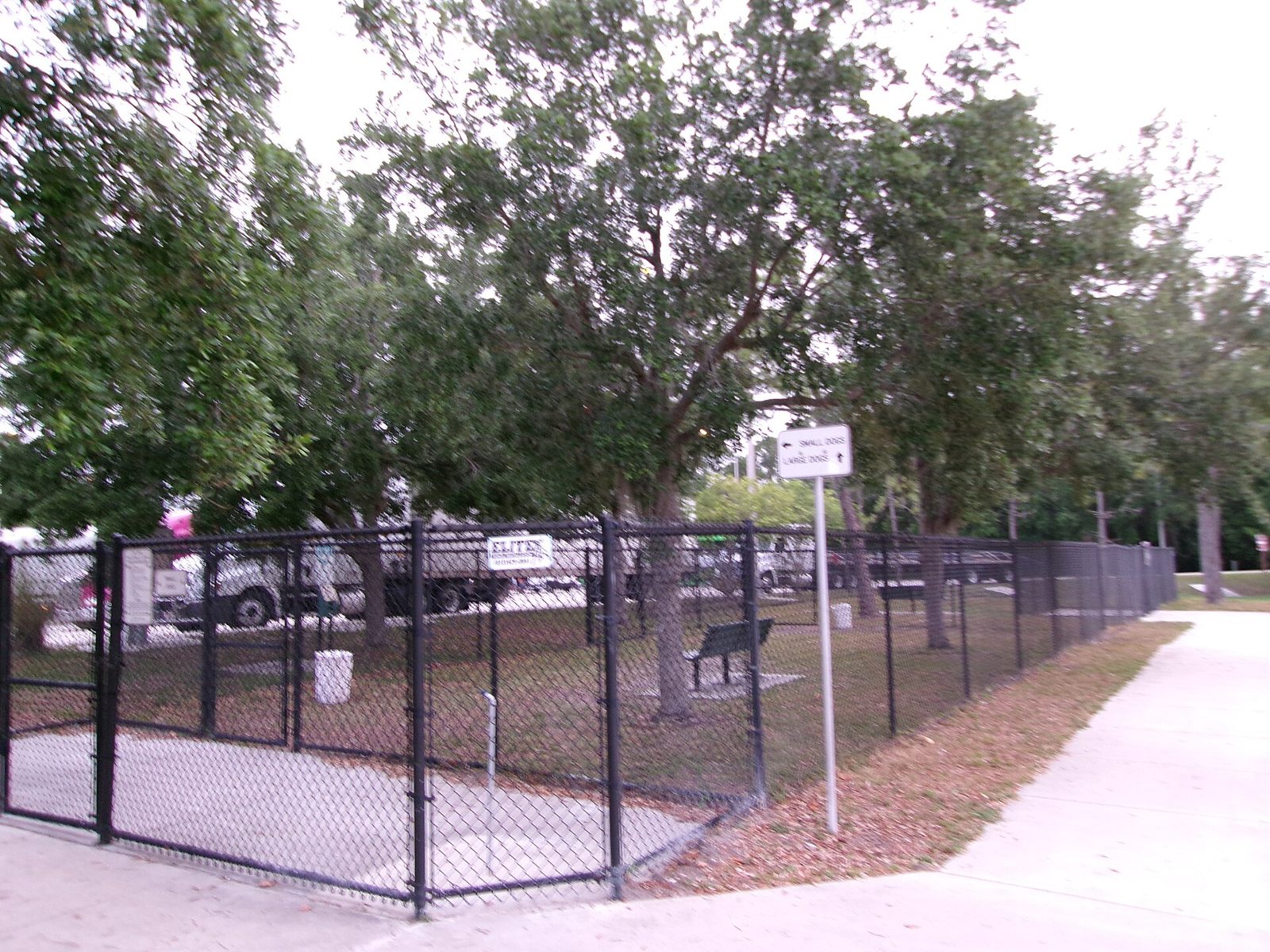 A fenced dog park with black chain-link fencing, benches, a trash can and a sign pointing to small dogs and large dogs, at a highway rest area under shade trees