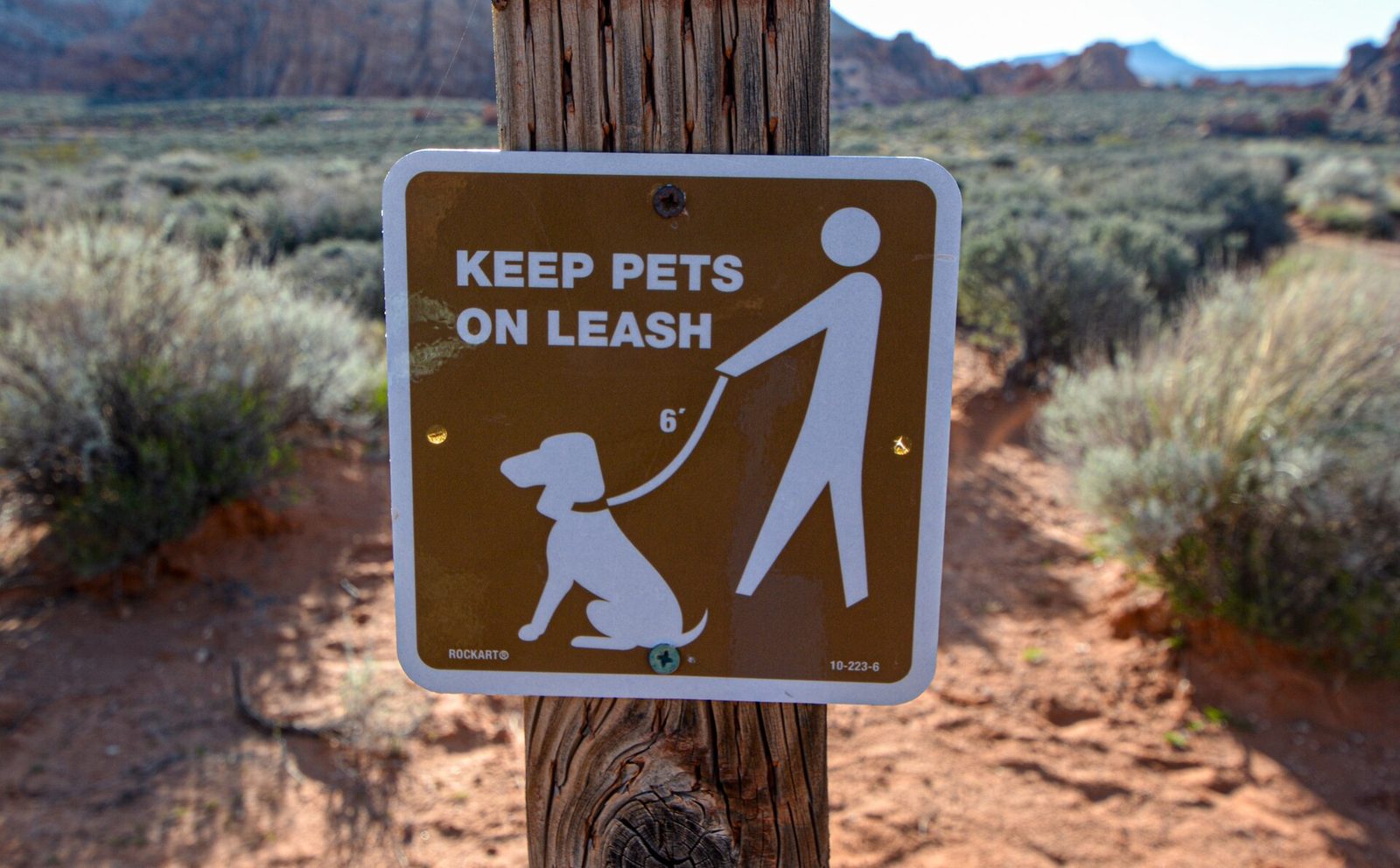 A brown trail sign on a wooden post reading Keep Pets On Leash, showing a walker with a dog on a 6-foot leash, in front of desert brush and red rock