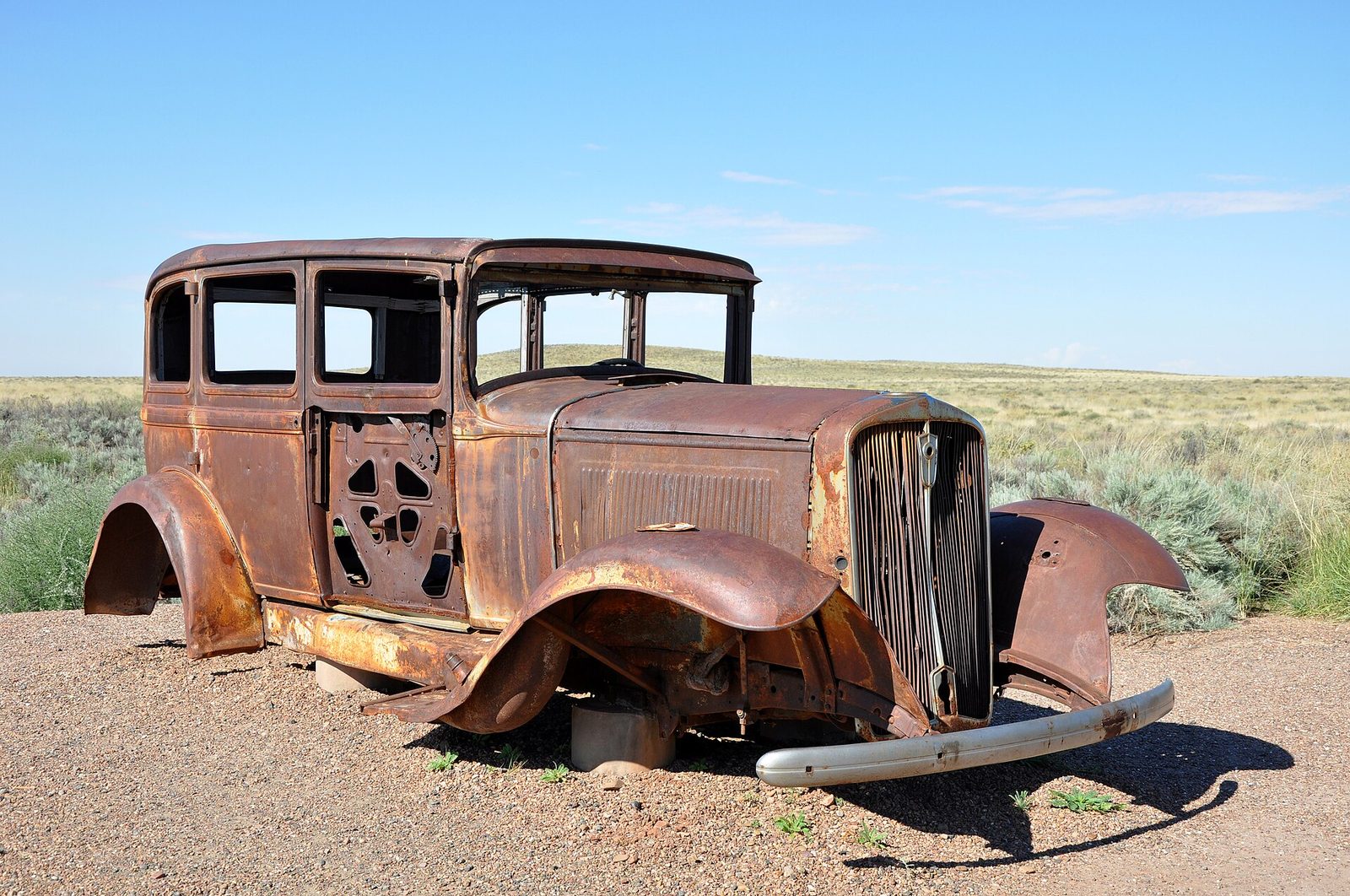 A rusted-out vintage Studebaker sedan sitting on gravel in front of open grassland under a blue sky
