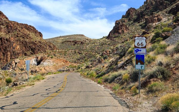 A narrow two-lane road climbing through rocky desert hills, with a brown Historic US 66 shield sign and America's Byways sign on the right