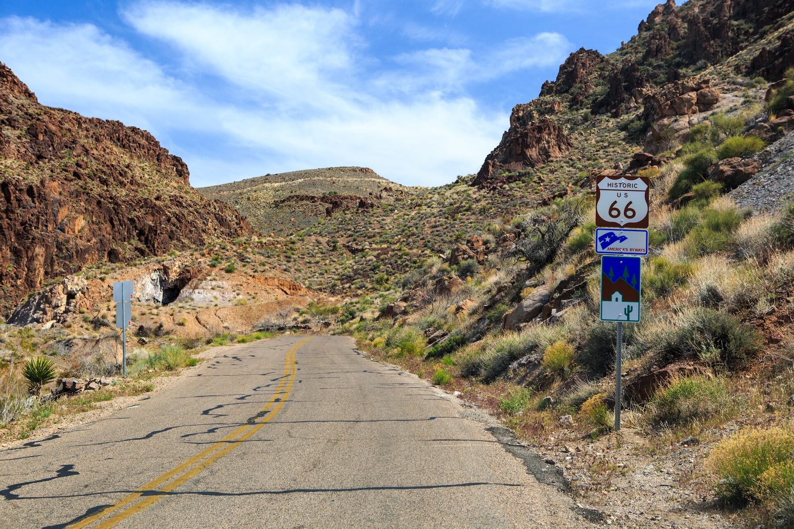 A narrow two-lane road climbing through rocky desert hills, with a brown Historic US 66 shield sign and America's Byways sign on the right