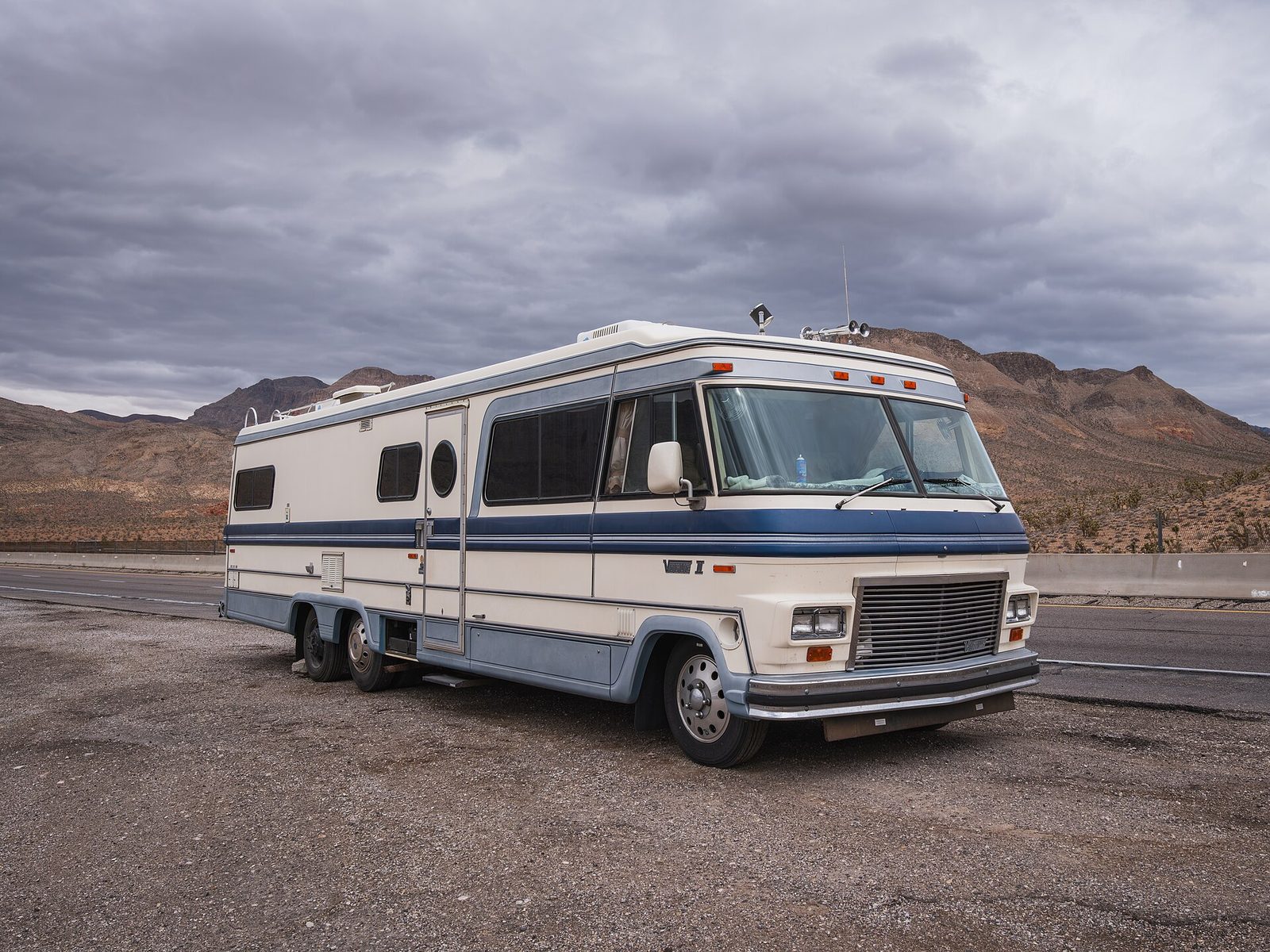 A vintage cream and blue Vogue motorhome parked on a gravel pull-out beside an interstate, with brown desert mountains and a cloudy sky behind it