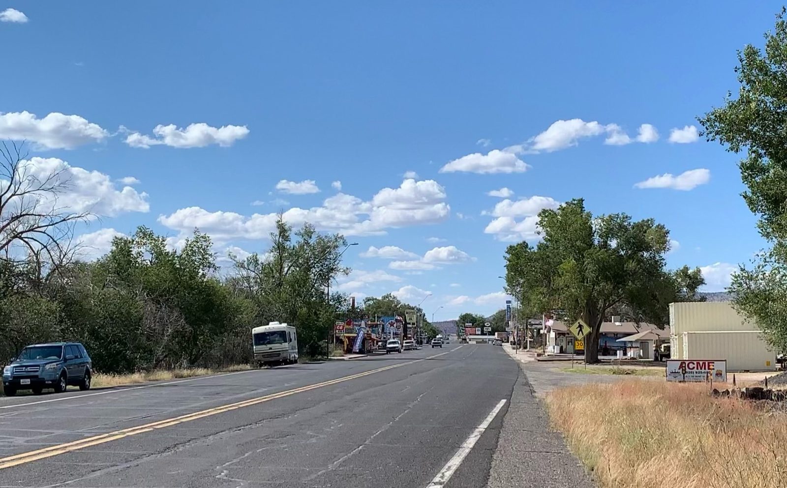 A wide two-lane main street in Seligman, Arizona, with a white motorhome parked on the left shoulder, colorful Route 66 souvenir shops in the distance and green trees on both sides