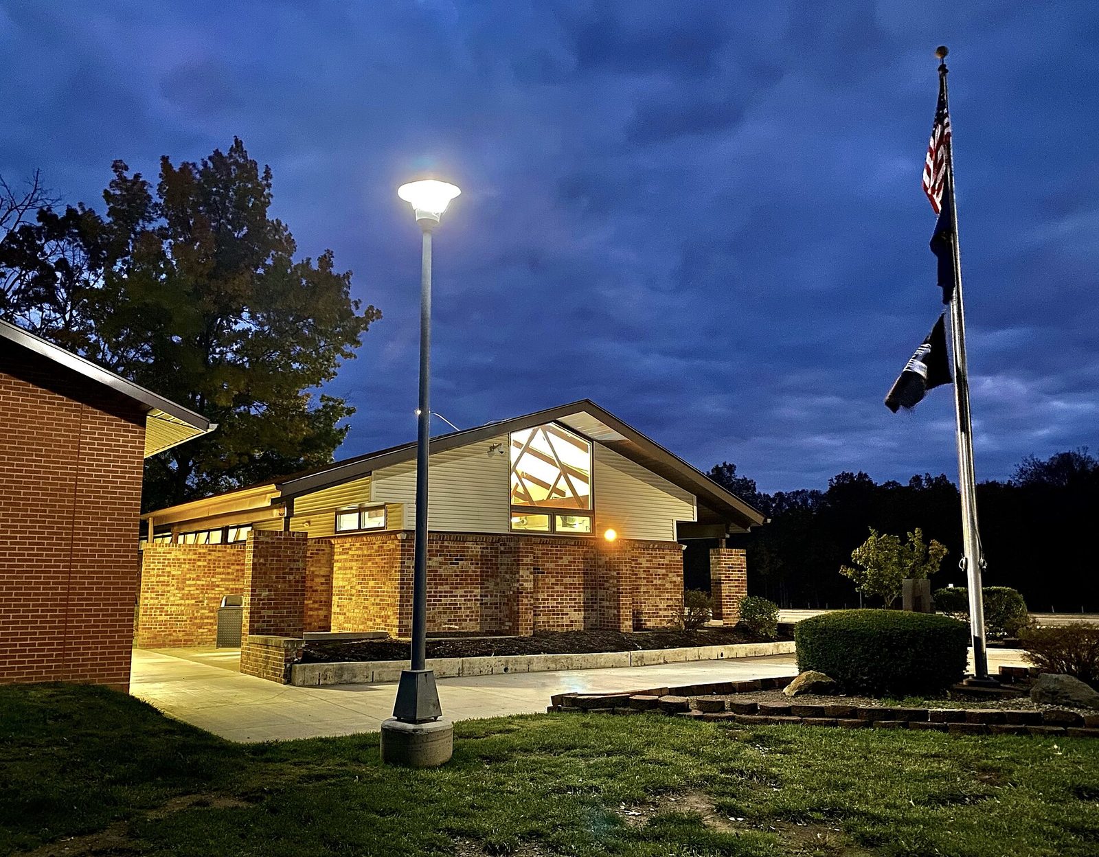 A brick highway rest area building at dusk with warm light glowing from its windows, a tall lit lamp post on the lawn in front and two flags on a pole under a dark blue cloudy sky