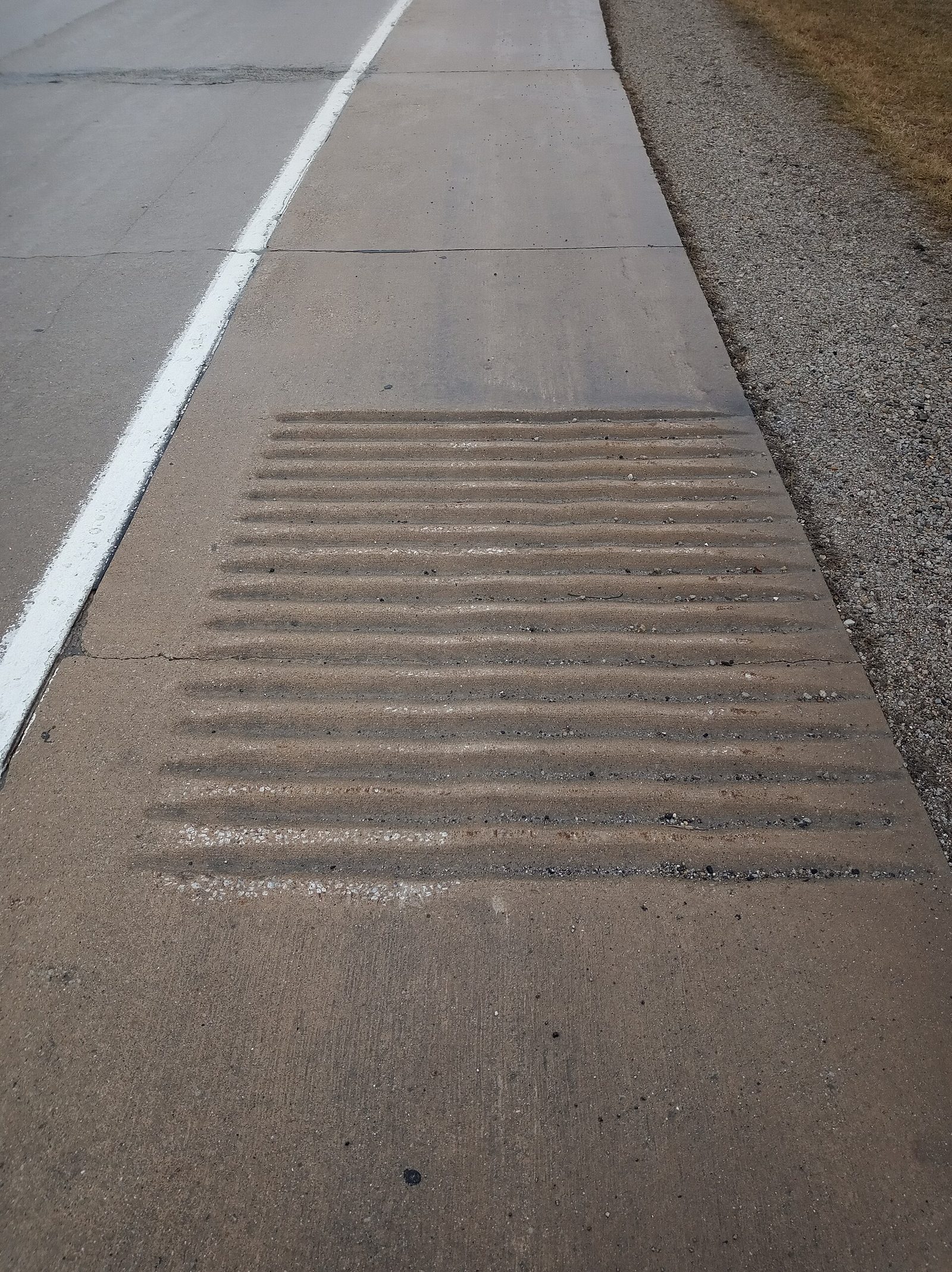 A close view of a concrete highway shoulder with a patch of grooved rumble strips beside the solid white edge line, with gravel and dry grass at the roadside
